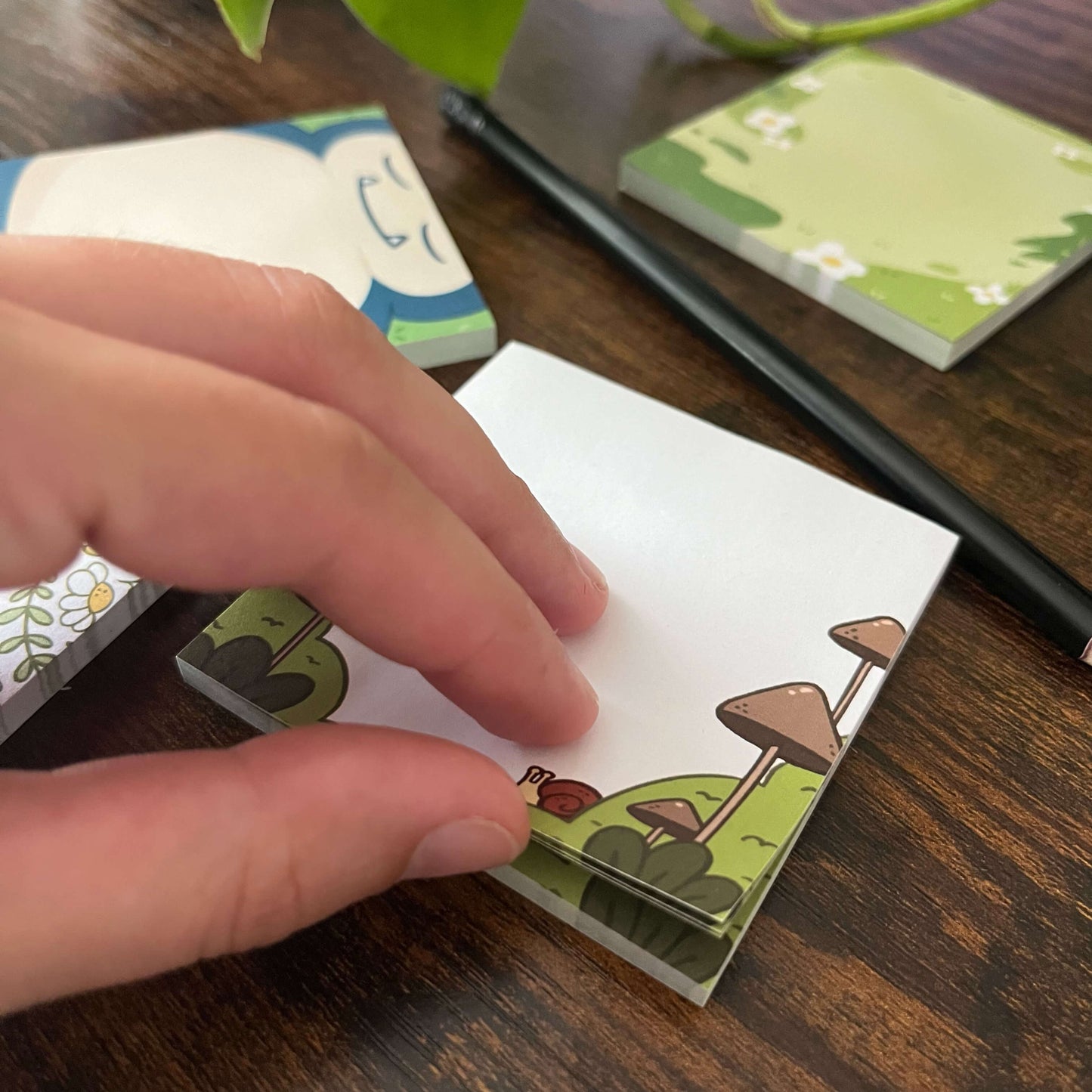 Decorative sticky notes of mushroom with snail being peeled. A pencil is nearby with two other sticky note pads.