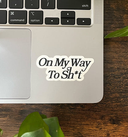 White sticker with the text "On My Way To Sh*t" written in elegant text, on the corner of a laptop.