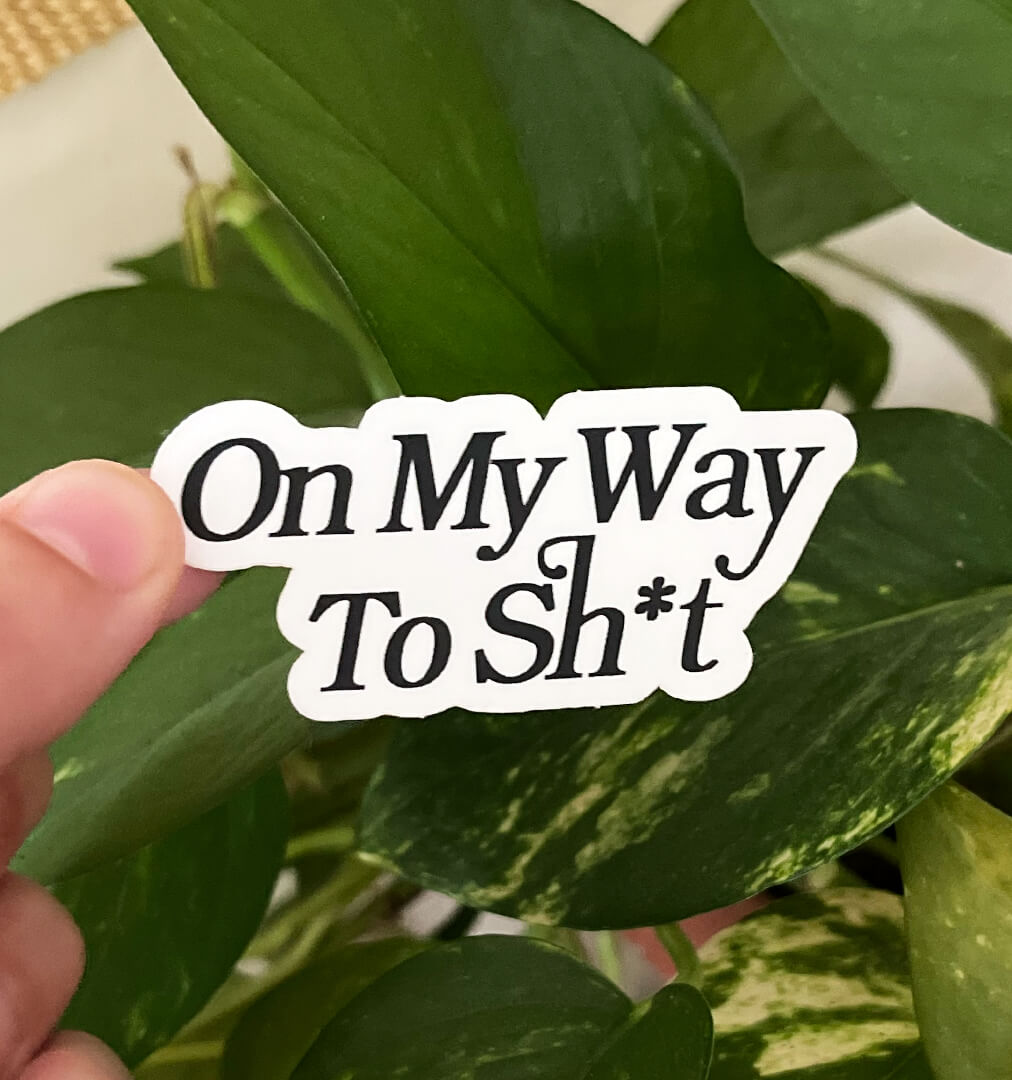 White sticker with the text "On My Way To Sh*t" written in elegant text, held by a hand in front of plants.