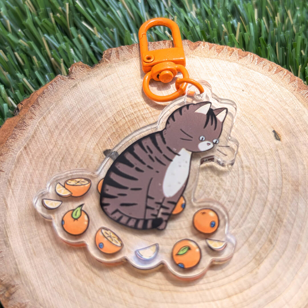 A clear acrylic keychain featuring a cartoon tabby cat surrounded by orange slices, attached to an orange lobster clasp, displayed on a wood slice with green grass in the background.