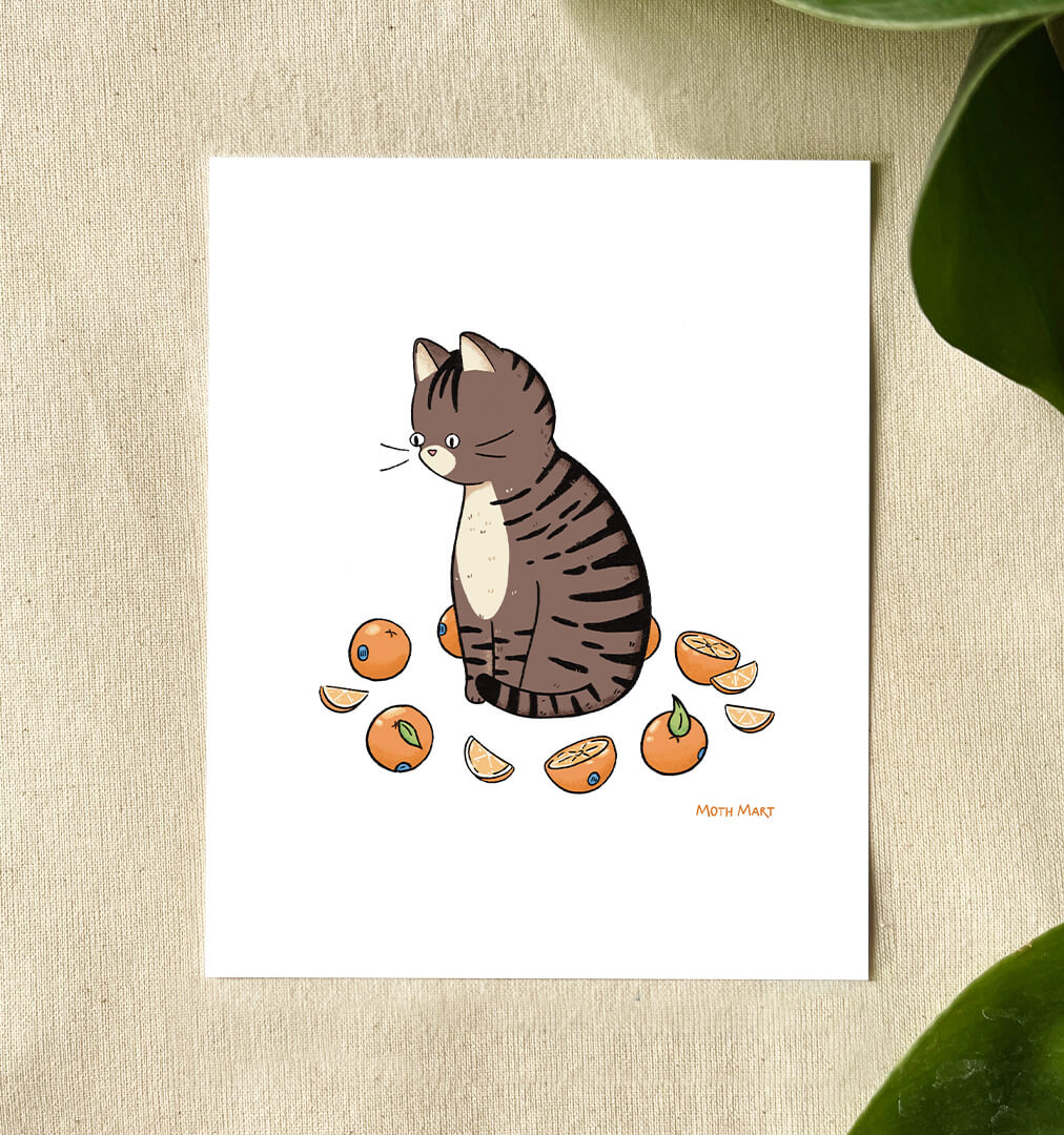 A white art print featuring a cute illustration of a brown tabby cat sitting among sliced oranges, displayed on a textured beige surface with green leaves in the background.