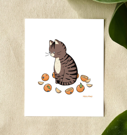 A white art print featuring a cute illustration of a brown tabby cat sitting among sliced oranges, displayed on a textured beige surface with green leaves in the background.