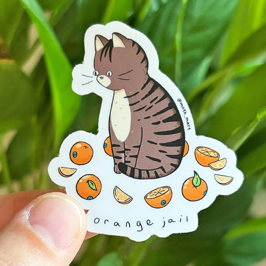 Die cut sticker of a brown tabby cat surrounded by oranges with the text "orange jail", being held in front of a leafy background.