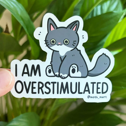 A die-cut sticker featuring a cartoon gray cat with the text 'I AM OVERSTIMULATED' and '@moth_mart', held by a hand in front of green plant leaves.