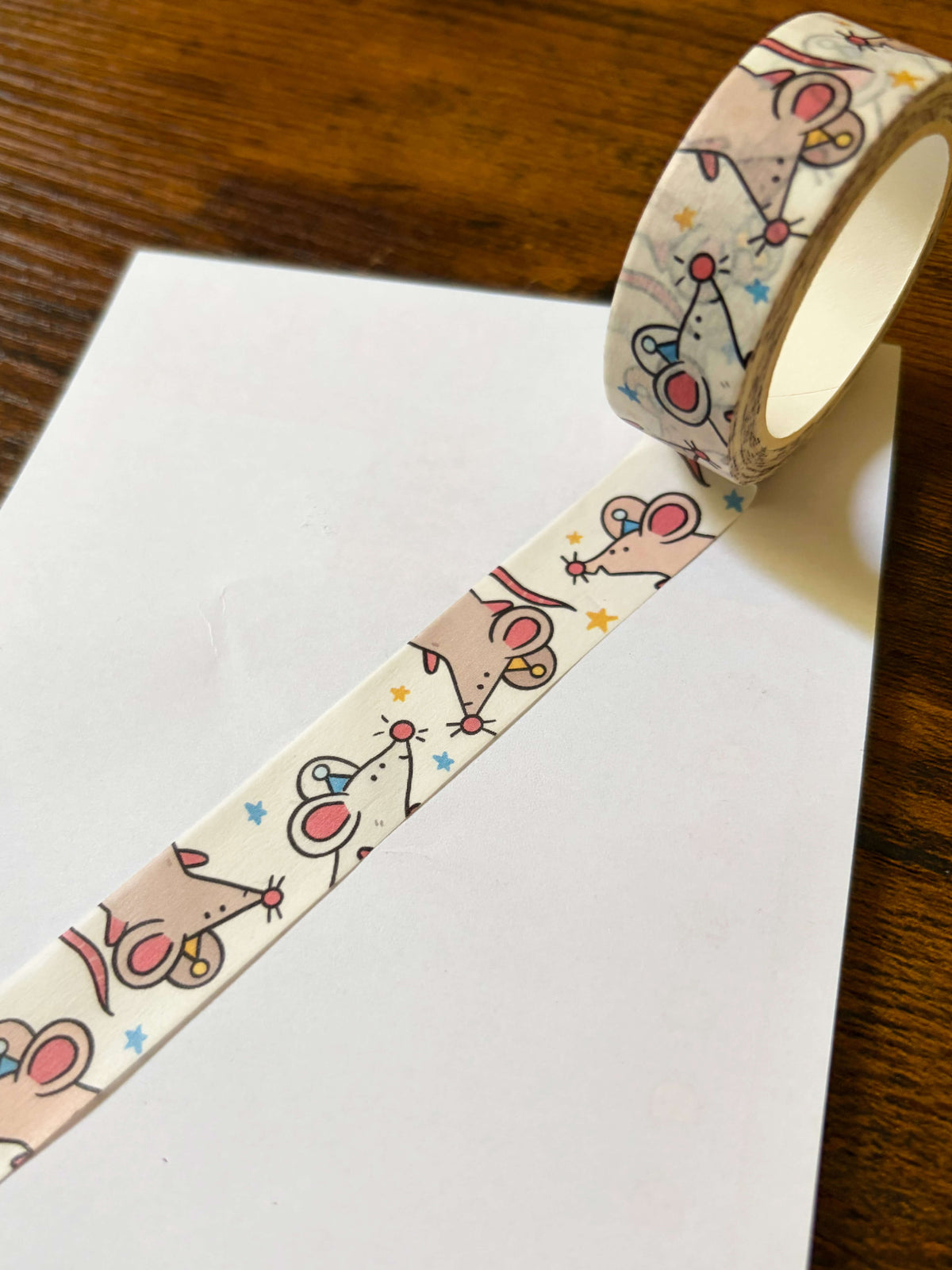 A roll of washi tape with a cute cartoon mouse pattern, partially unrolled over a white sheet of paper on a wooden table.