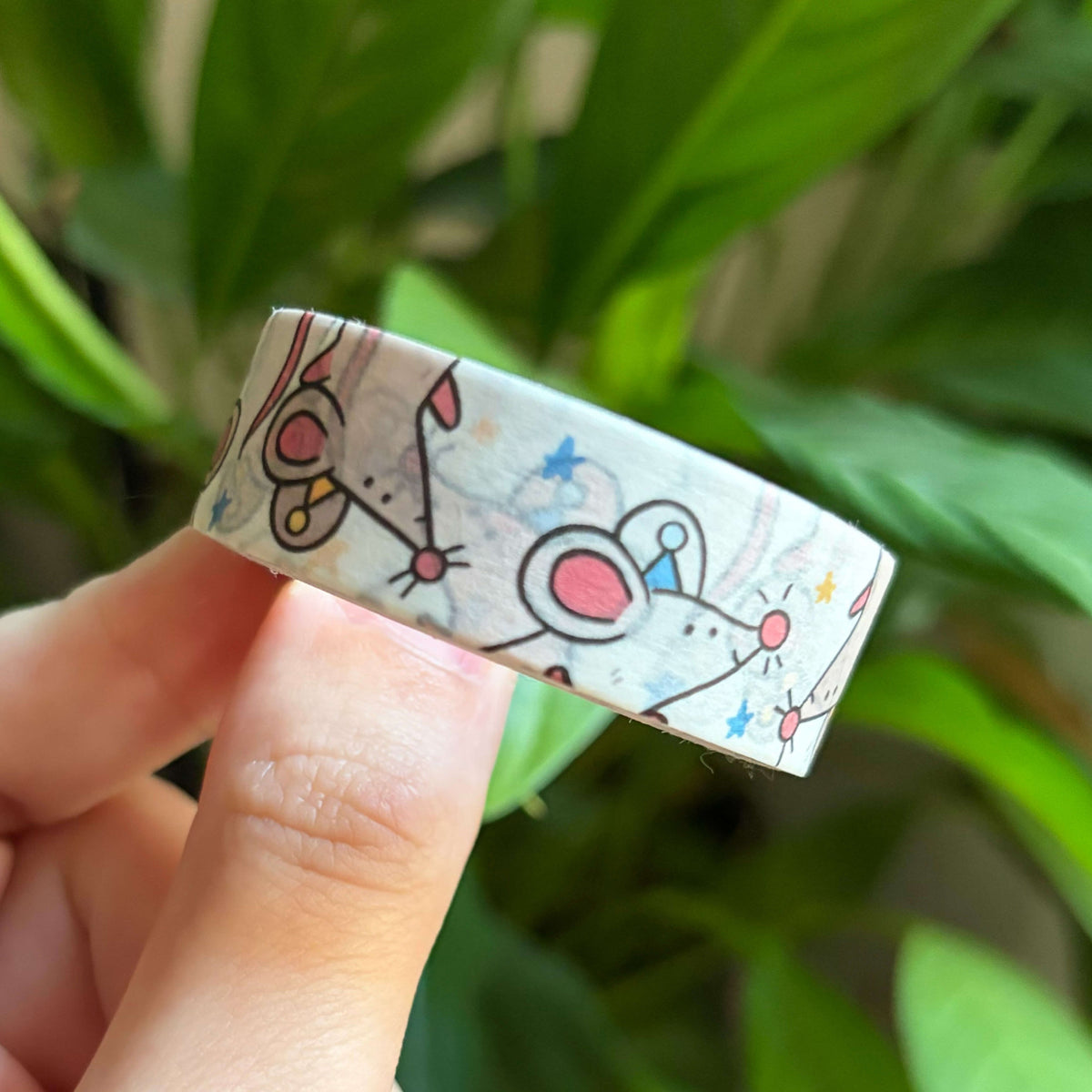 A hand holding a roll of washi tape with a cartoon mouse pattern against a background of green houseplants.