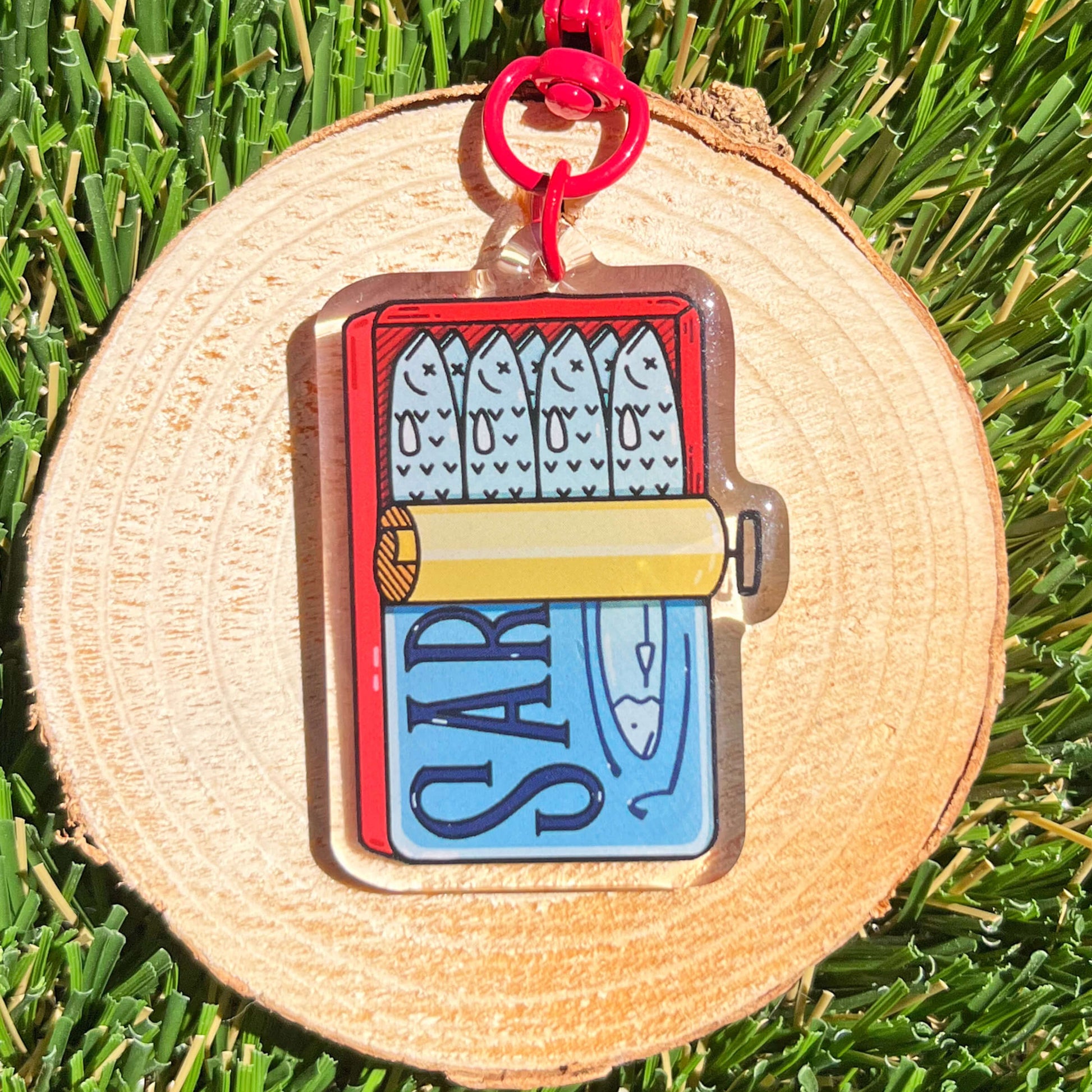 Keychain with illustration of canned sardines with a red hook, sitting on wood background with grass.