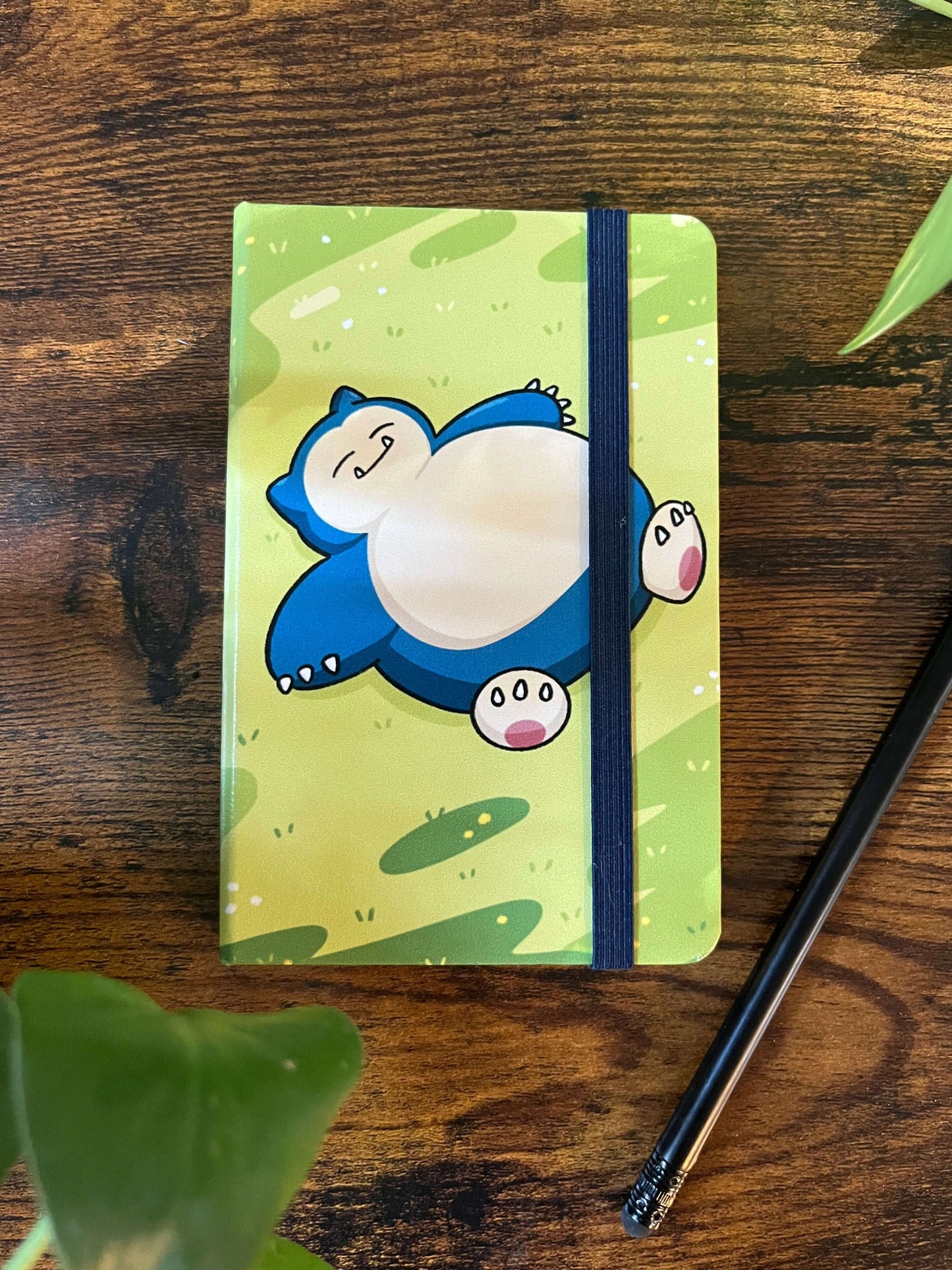 A notebook on a wood table with a blue strap holding it shut. Notebook cover depicts a sleeping, large, blue creature.
