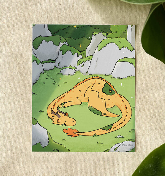 A vertical art print featuring a whimsical illustration of a sleeping orange dragon curled up in a grassy green landscape with gray rocks, displayed on a textured beige fabric background with green leaves in the corners.
