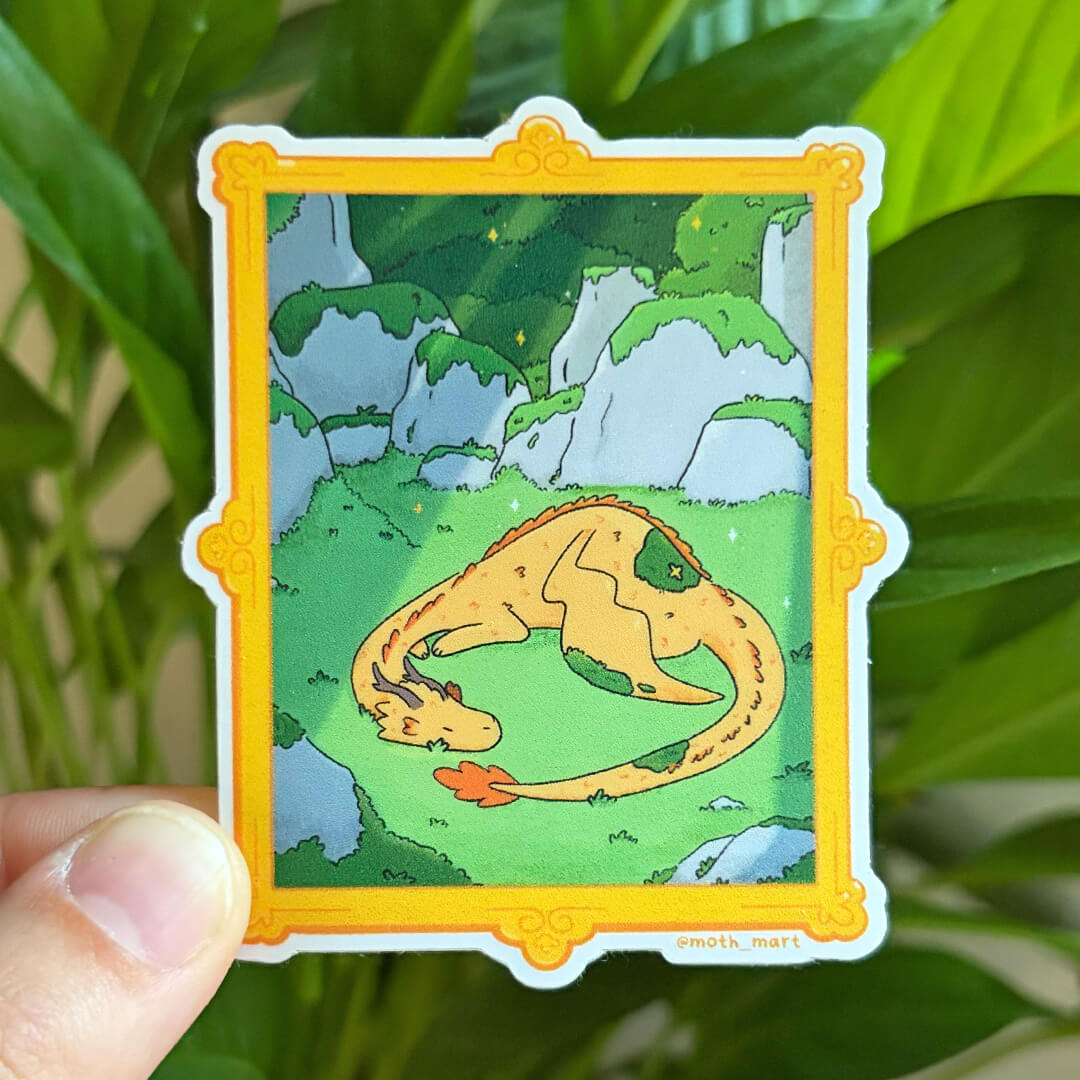 A hand holds a die-cut sticker featuring a cartoon illustration of a yellow dragon curled up on a green landscape with rocks, framed by a yellow border. The sticker is in front of green plant leaves.