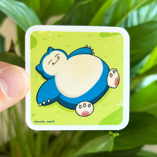 Sticker of a sleeping blue creature on grass, held against a blurred houseplant background. 