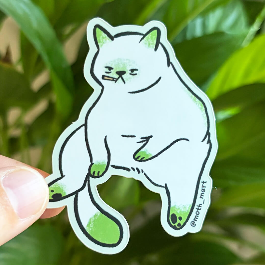 A die-cut sticker featuring a smoking cartoon white cat with green paws and ears, held by a person's fingers in front of a blurred green plant background.