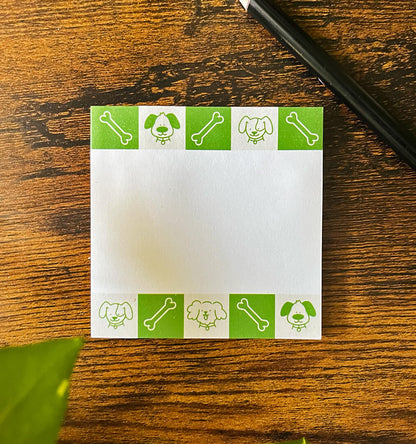 Sticky notepad on wood background depicting green and beige squares featuring cartoon dogs and bones. Pencil off to the side.