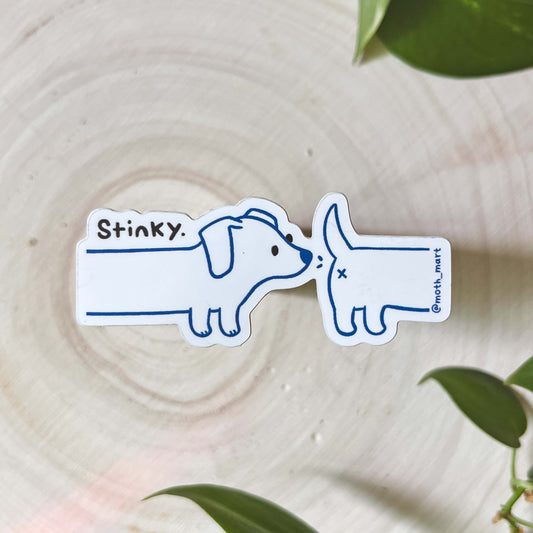 Sticker of a dog sniffing another dog's butt with 'Stinky' text on a wooden surface with green leaves.