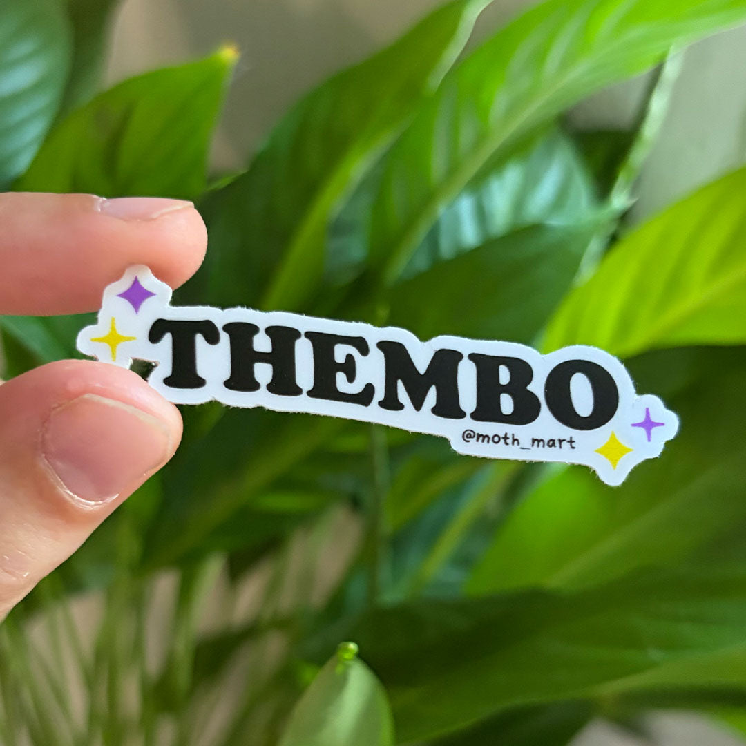 A hand holds a die-cut sticker that reads “THEMBO” in bold black letters on a white background with small purple and yellow star accents. The sticker also includes the text “@moth_mart."