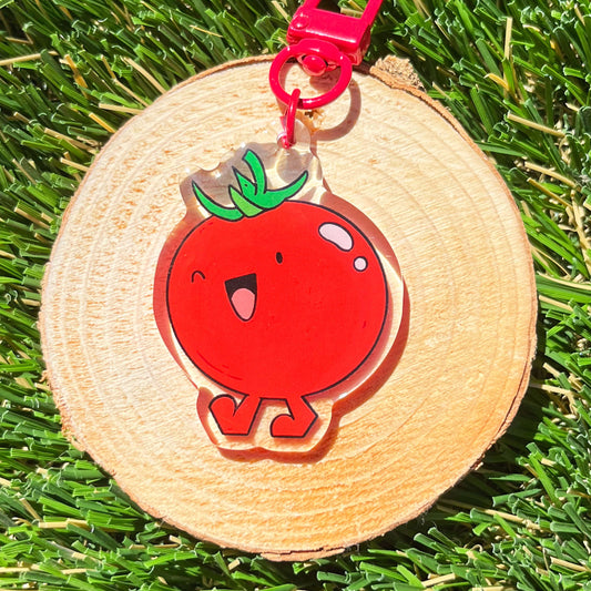 Red cartoon tomato keychain on a wooden disc with a pink clip, placed on grass.