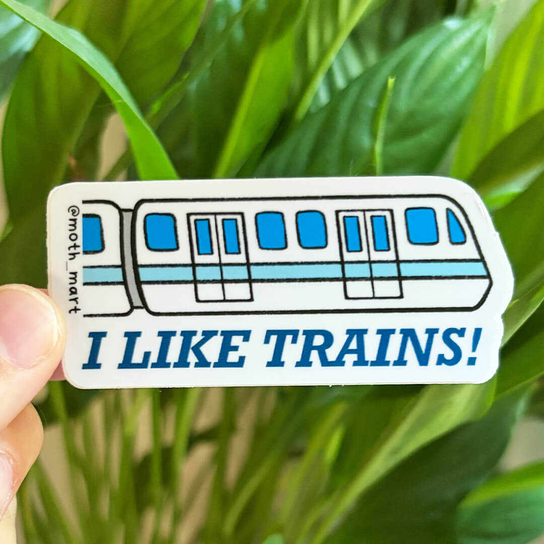 Die cut sticker depicting a blie and white train with the text "I like trains!" held by a hand in front of a background of houseplants.