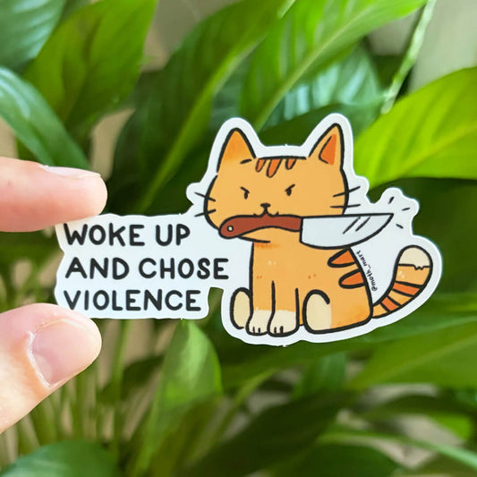 A hand holds a die-cut sticker featuring an orange tabby cartoon cat holding a knife in its mouth, with the text 'WOKE UP AND CHOSE VIOLENCE' on a white background. The sticker is photographed in front of blurred green houseplant leaves.