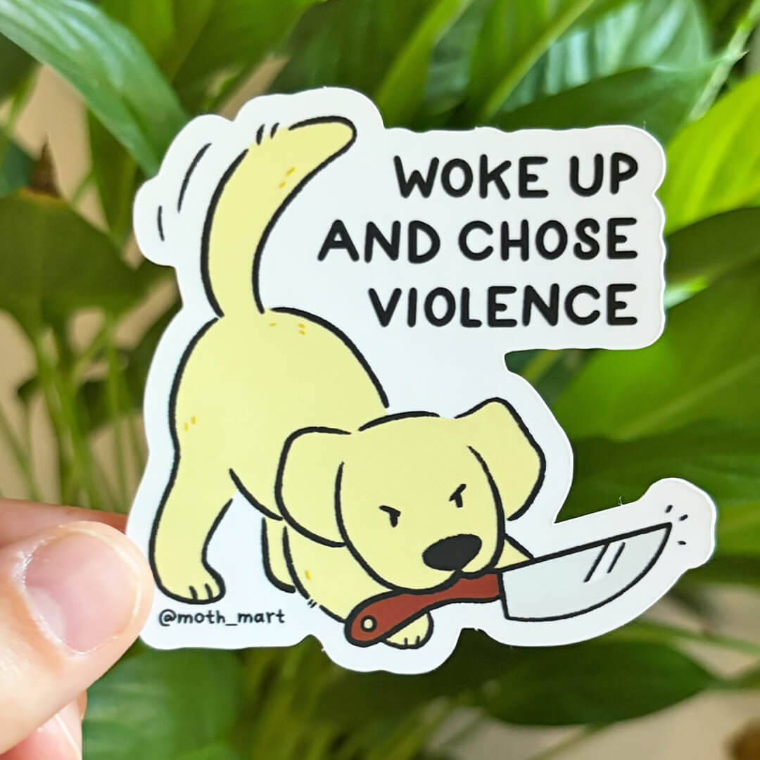 A die-cut sticker held by a hand against a background of green houseplants. The sticker features a cartoon yellow puppy biting a knife and has the text 'WOKE UP AND CHOSE VIOLENCE' in black capital letters.