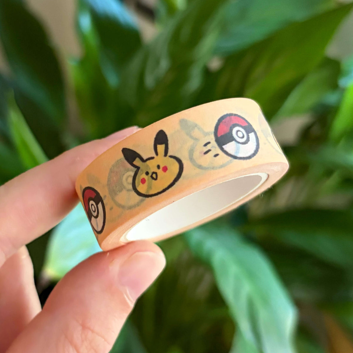 A hand holding a roll of washi tape featuring a yellow creature and red ball designs, with a blurred background of green houseplants.
