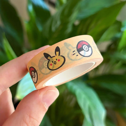 A hand holding a roll of washi tape featuring a yellow creature and red ball designs, with a blurred background of green houseplants.