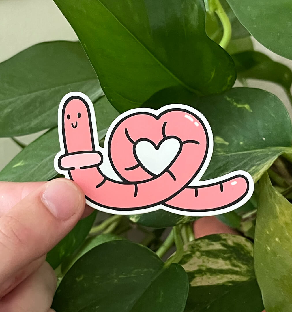 Sticker of a pink worm in a heart shape held by a hand in front of green plants.