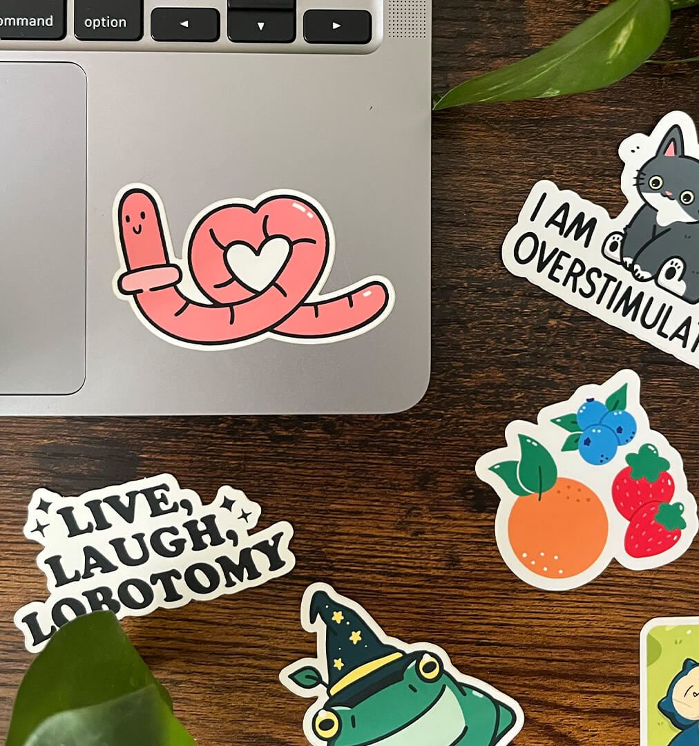 Sticker of a pink worm in a heart shape on the corner of a laptop, surrounded by 5 other stickers. 